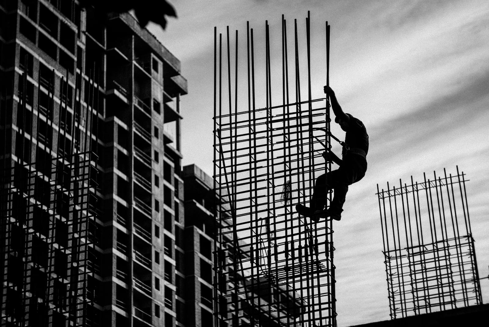 Somos Albañileria El Mghari, tu aliado de confianza en Murcia. Ofrecemos servicios de albañilería de alta calidad Black and white silhouette of a construction worker climbing steel rods on a building site.